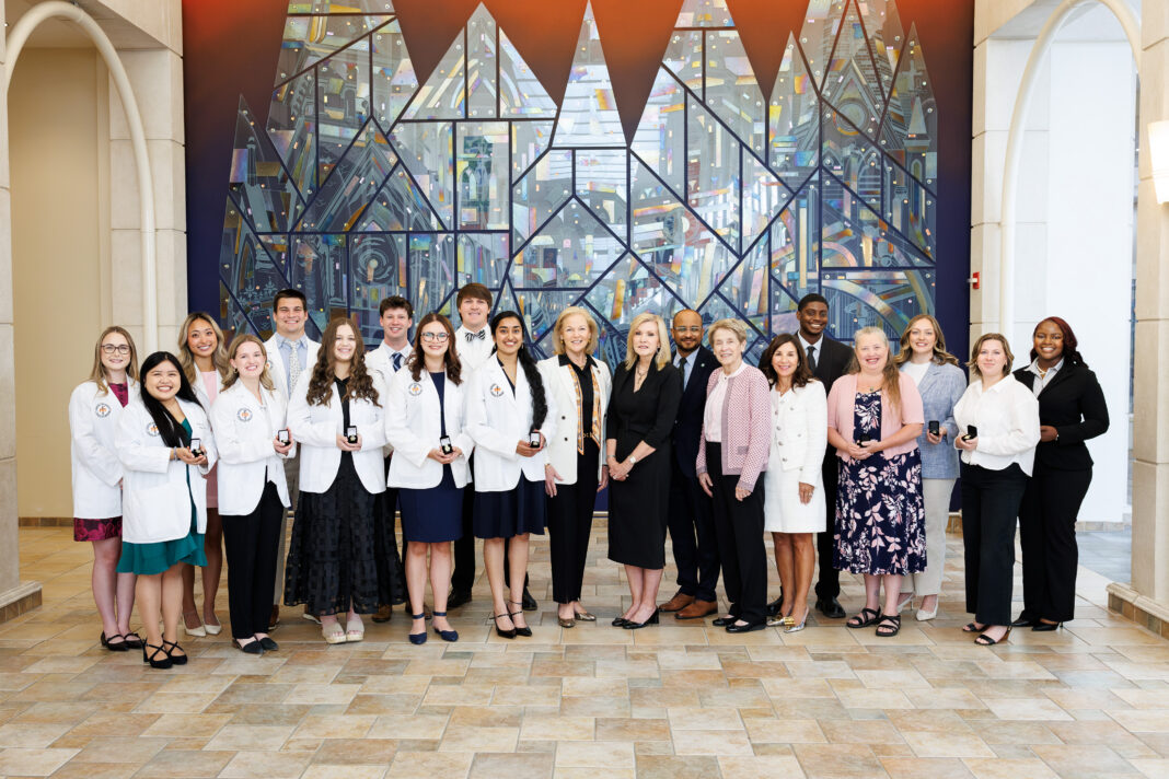 A group of people, some in white coats, stand together indoors in front of a large, geometric stained glass mural.