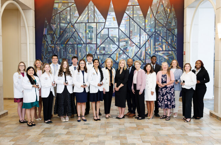 A group of people, some in white coats, stand together indoors in front of a large, geometric stained glass mural.