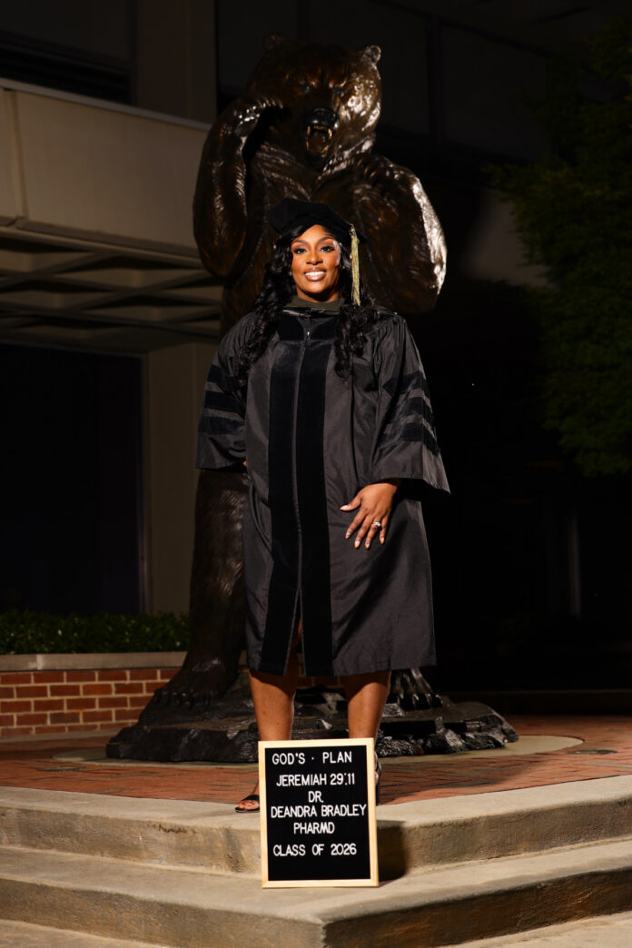 Deandra Bradley, wearing graduation attire, stands in front of a bear statue.
