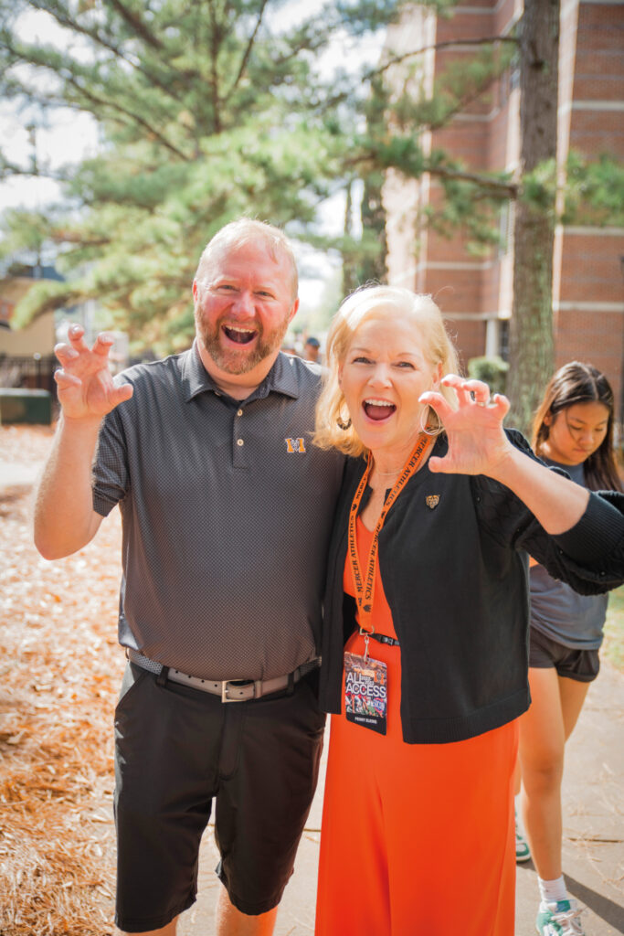 Jason Elkins and President Dr. Penny L. Elkins pose outdoors, smiling and making playful claw gestures, with trees and a building in the background.