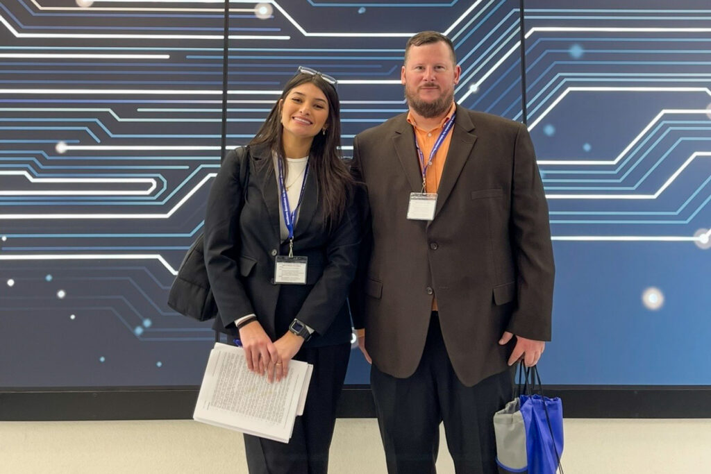 Frida Danson and Dr. Johnathan Yerby stand in front of a blue backdrop with circuit-like patterns, both wearing conference badges.