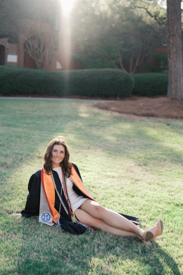 Gracie Wood in graduation attire sits on grass in sunlight, smiling at the camera with trees and buildings in the background.