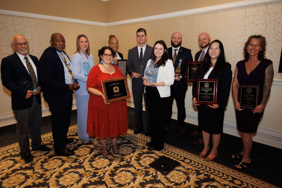 Ten people stand in a room holding plaques and trophies, posing for an award ceremony group photo.