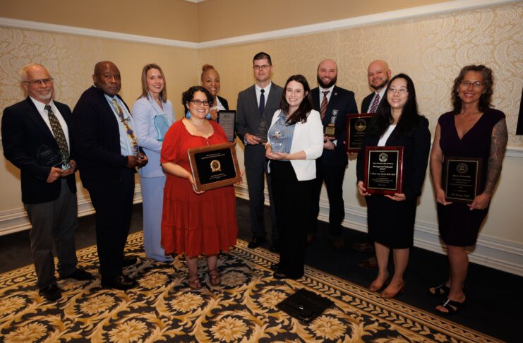Ten people stand in a room holding plaques and trophies, posing for an award ceremony group photo.