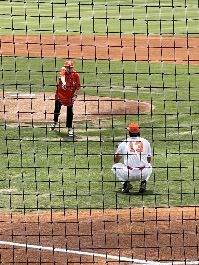 Dr. Mary Wilder throws a baseball pitch to a crouching catcher on a baseball field behind a protective net.