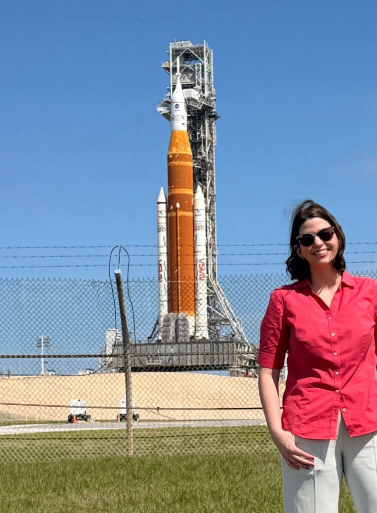 Katie Konans stands in front of a fenced area with a NASA rocket on a launch pad in the background.