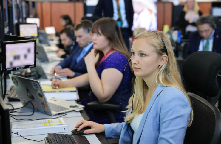 Several people sit at desks working on computers in an office setting, focused on their monitors.