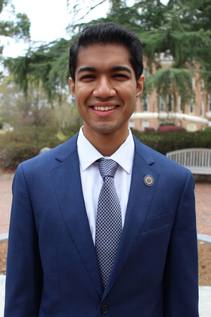 A young man in a blue suit and tie stands outside, smiling at the camera with trees and a building in the background.