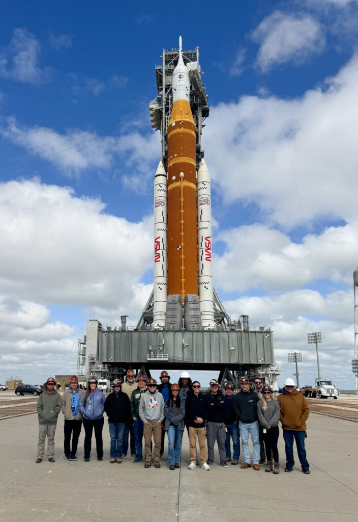 A group of people stand in front of a NASA rocket on a launch pad under a partly cloudy sky.