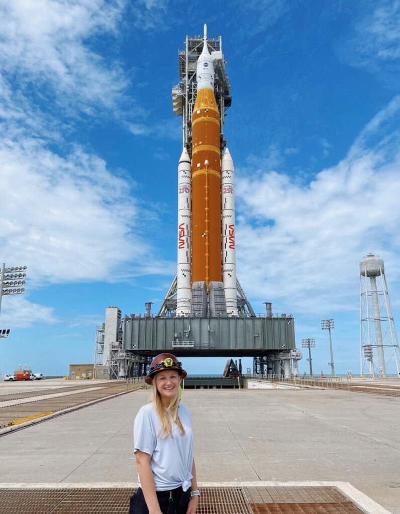 A woman stands smiling in front of a NASA rocket on a launch pad under a partly cloudy sky.