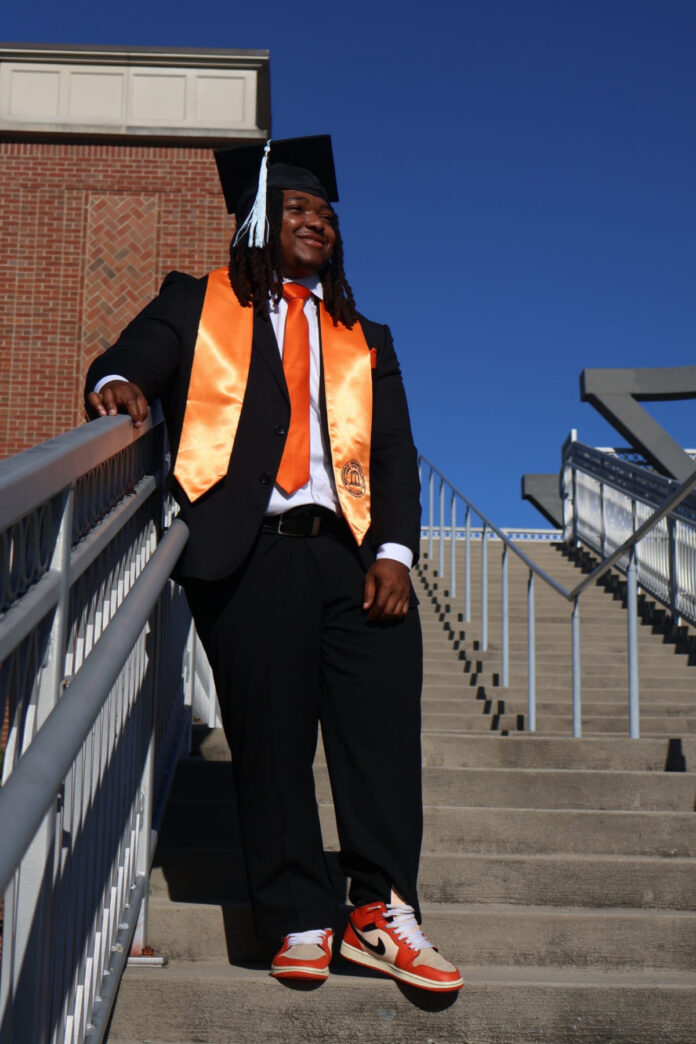 Nicholas Milsap in cap and gown stands on outdoor steps, wearing an orange sash and matching tie and sneakers.