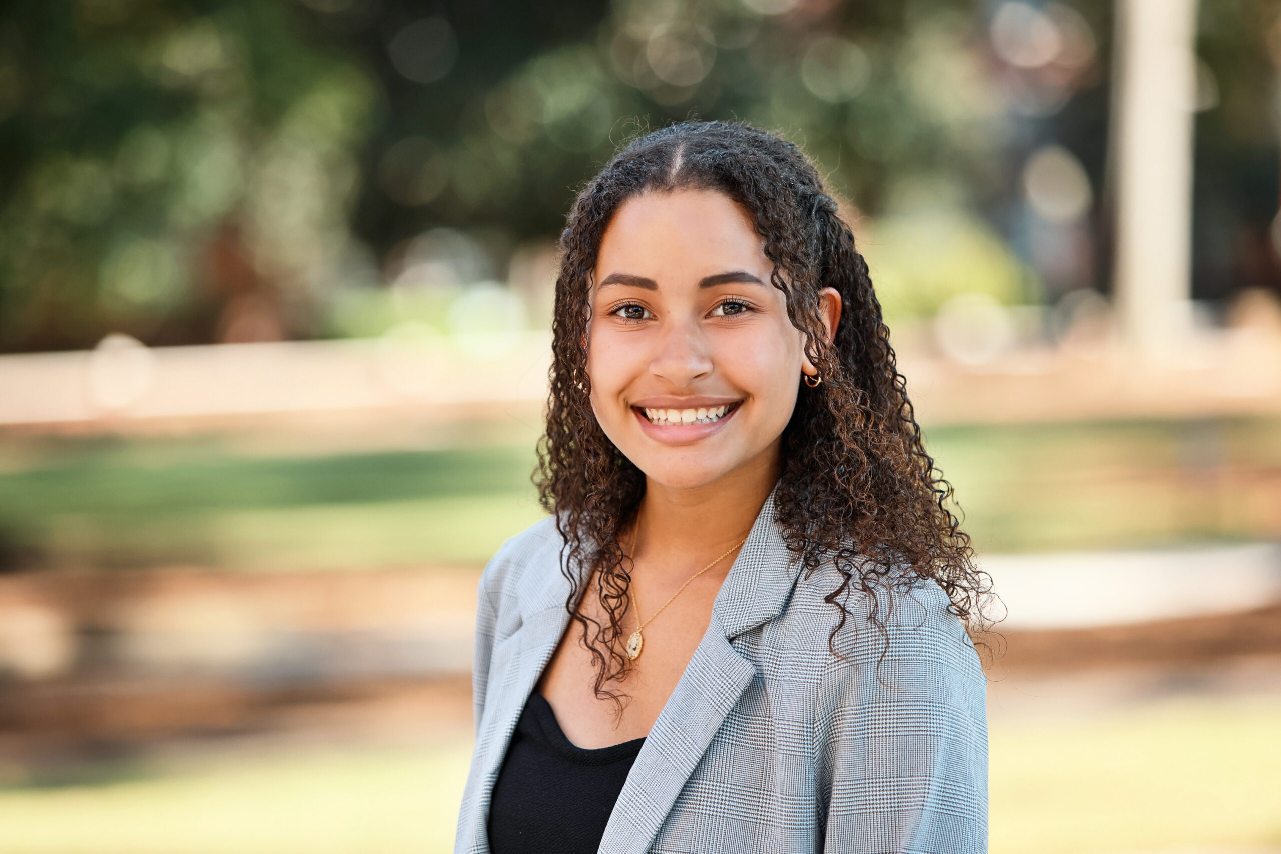 A woman with curly hair in a gray blazer smiles outdoors with green trees and sunlight in the background.