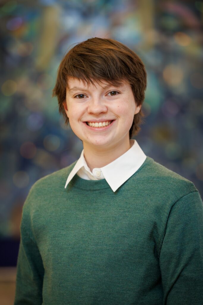 A person with short brown hair, wearing a green sweater over a white collared shirt, smiling at the camera.