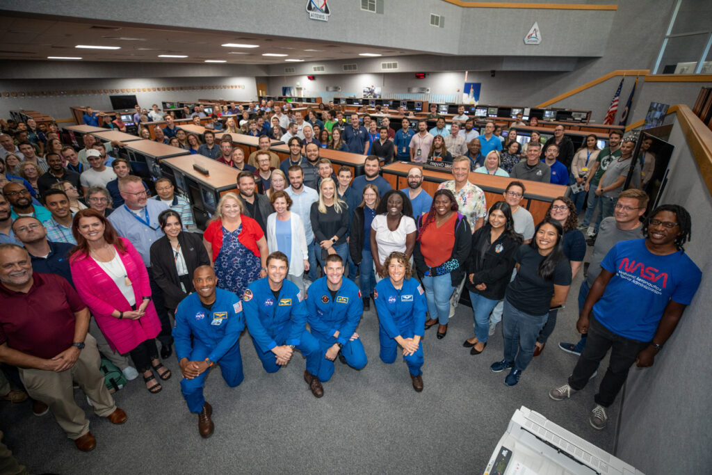 A large group of people, including astronauts in blue suits, pose for a group photo in a NASA mission control room.