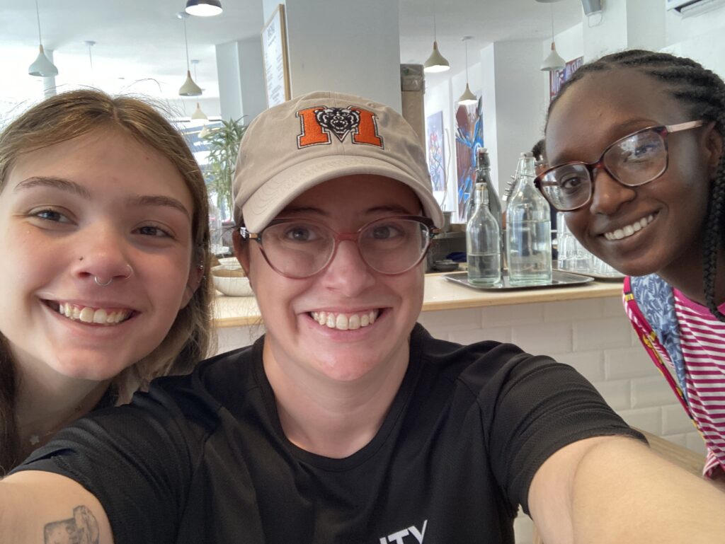 Three people smiling and posing for a selfie in a bright indoor setting with glass bottles on the counter behind them.