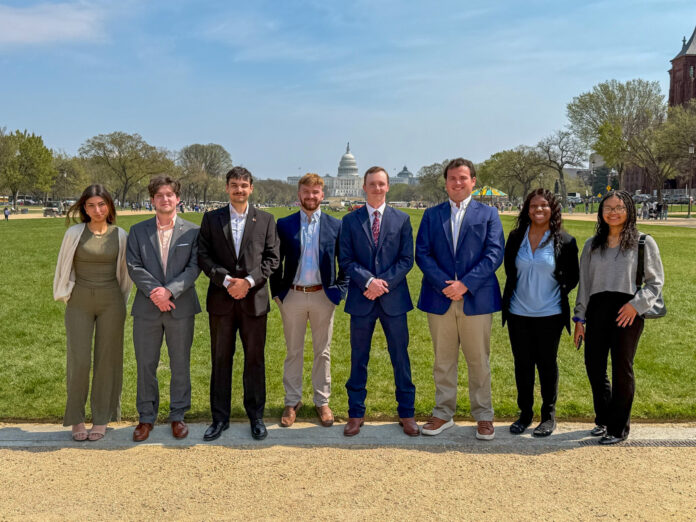Eight people stand in a row outside on a path with the U.S. Capitol building visible in the background.