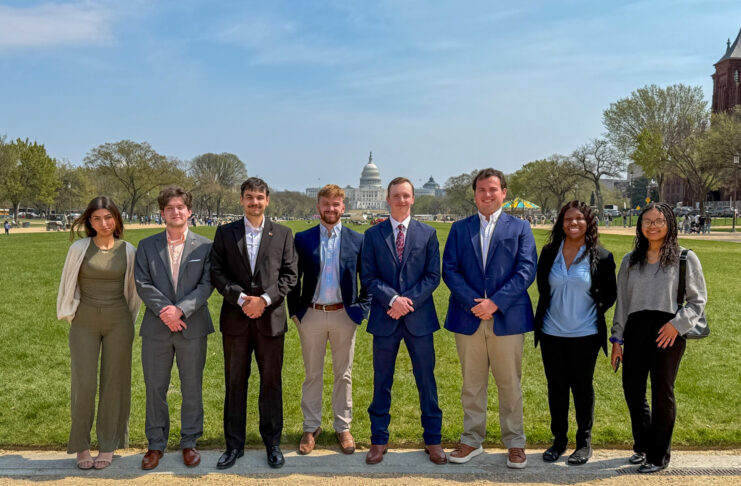 Eight people stand in a row outside on a path with the U.S. Capitol building visible in the background.