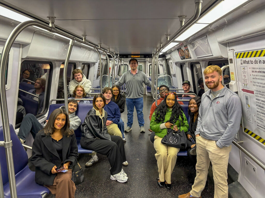 A group of people sit and stand inside a subway train car, facing the camera and smiling.