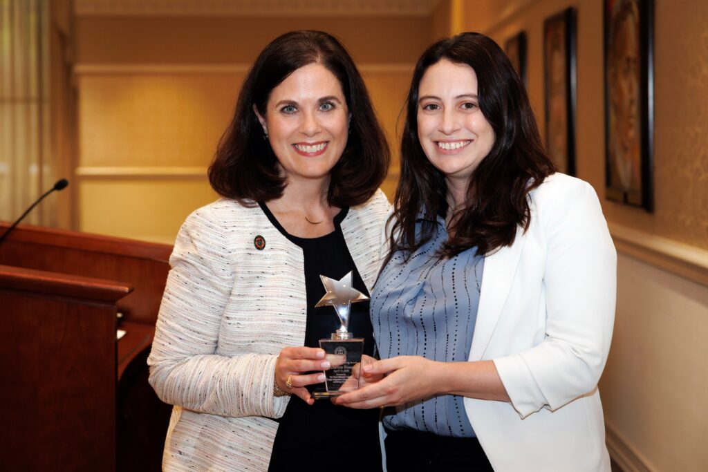 Two women standing indoors, smiling, and holding a glass trophy with a silver star on top.