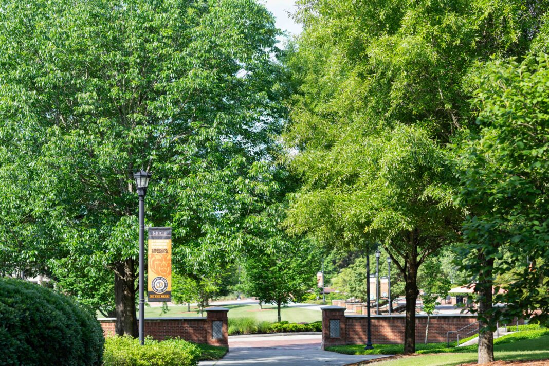 Pathway surrounded by green trees, brick columns, and a campus banner on a sunny day.