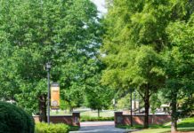 Faculty and Staff Notables | May 2026 Pathway surrounded by green trees, brick columns, and a campus banner on a sunny day.