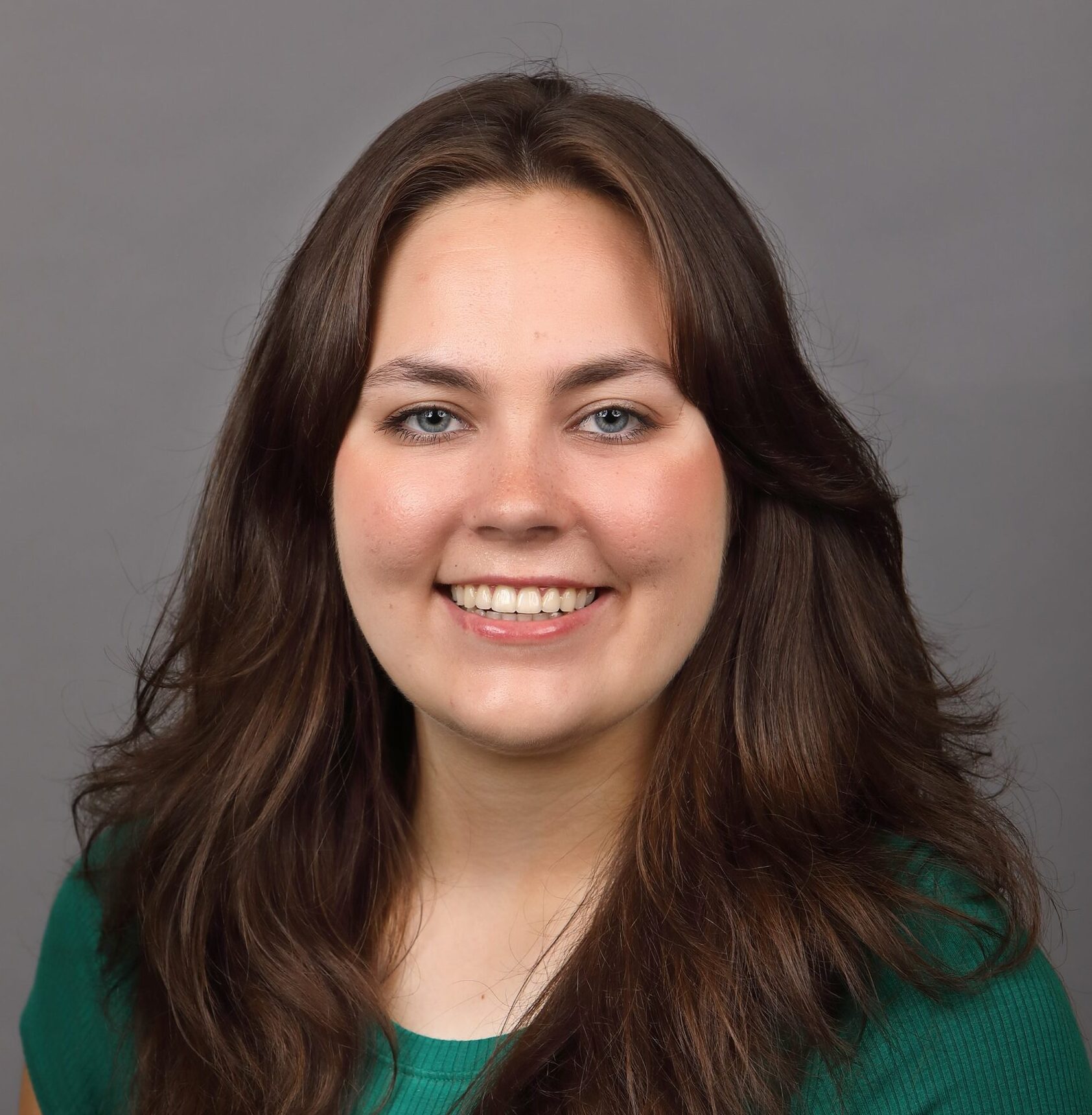 Young woman with long brown hair wearing a green top, smiling at the camera against a plain gray background.