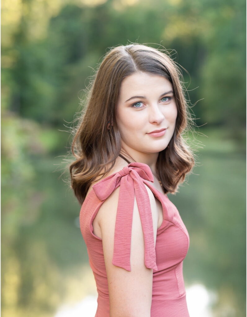 Young woman with light brown hair in a pink dress stands outdoors near water with a blurred green background.
