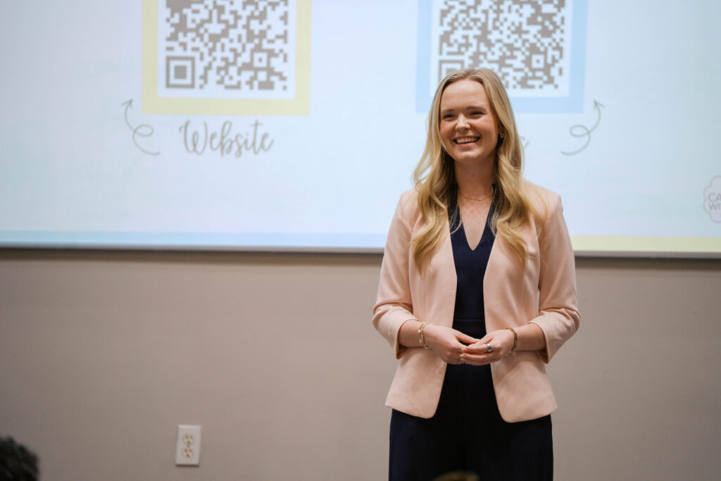 Woman in business attire giving a presentation, standing in front of a screen displaying QR codes and the word “Website.”.