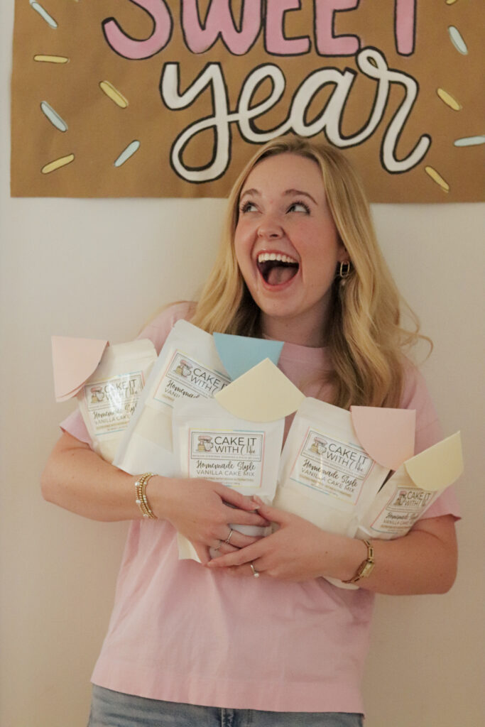 Woman holding several cake mix packages, smiling in front of a sweet year sign on the wall.