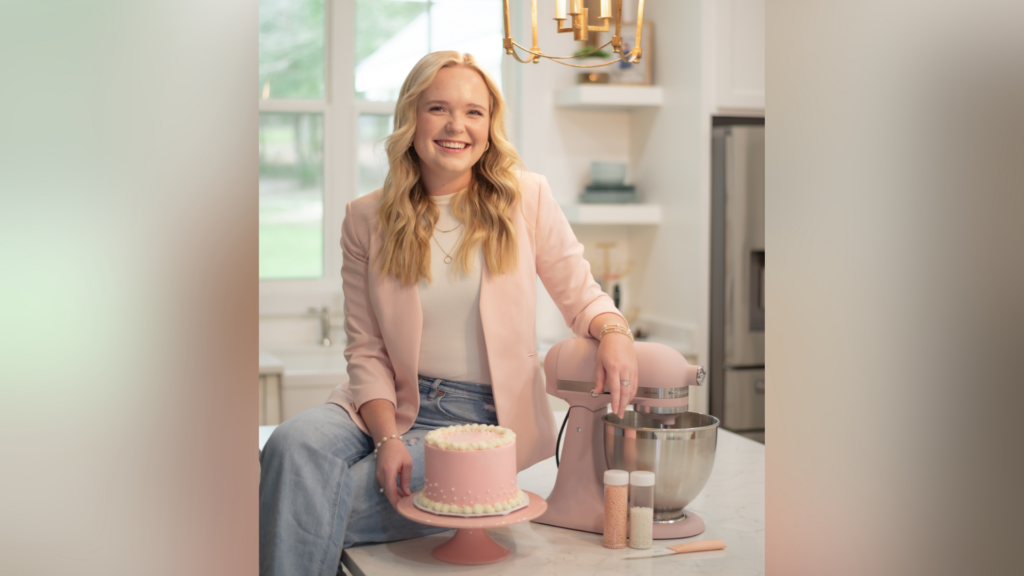 Woman sitting on a kitchen counter with a pink frosted cake, a pink mixer, and sprinkles in a modern kitchen.