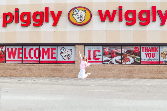 Person jumps in front of a Piggly Wiggly grocery store with large red signage and welcome banners on the windows.
