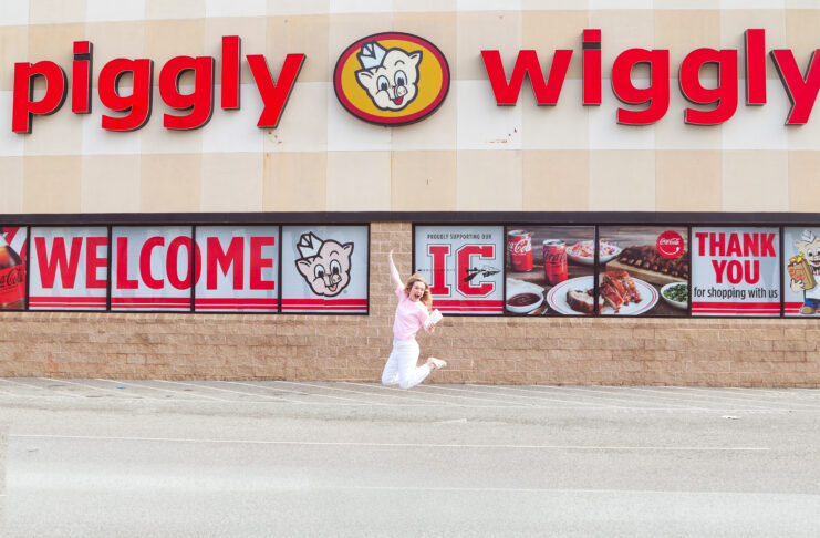Person jumps in front of a Piggly Wiggly grocery store with large red signage and welcome banners on the windows.