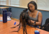 Hair care club encourages students to celebrate their natural hair A woman sits at a table braiding hair on a mannequin head, with tools and a water bottle nearby.