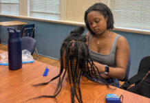 Hair care club encourages students to celebrate their natural hair A woman sits at a table braiding hair on a mannequin head, with tools and a water bottle nearby.