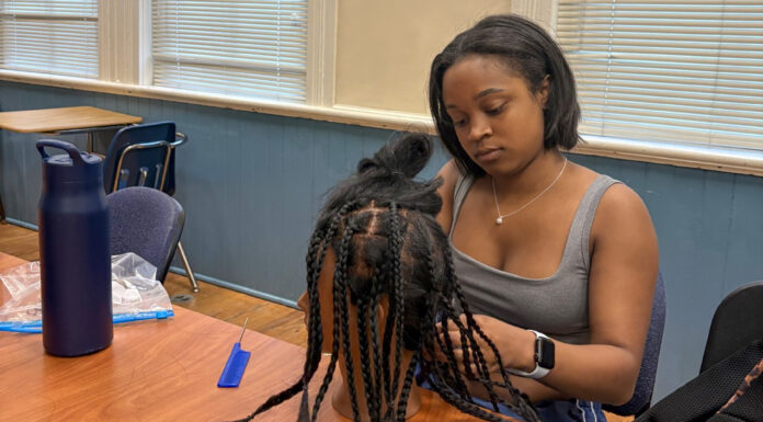 Hair care club encourages students to celebrate their natural hair A woman sits at a table braiding hair on a mannequin head, with tools and a water bottle nearby.