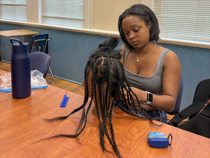 A woman sits at a table braiding hair on a mannequin head, with tools and a water bottle nearby.