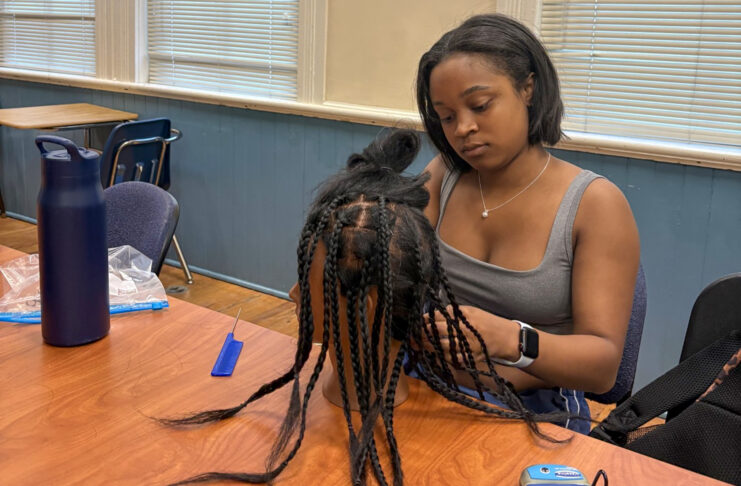 A woman sits at a table braiding hair on a mannequin head, with tools and a water bottle nearby.