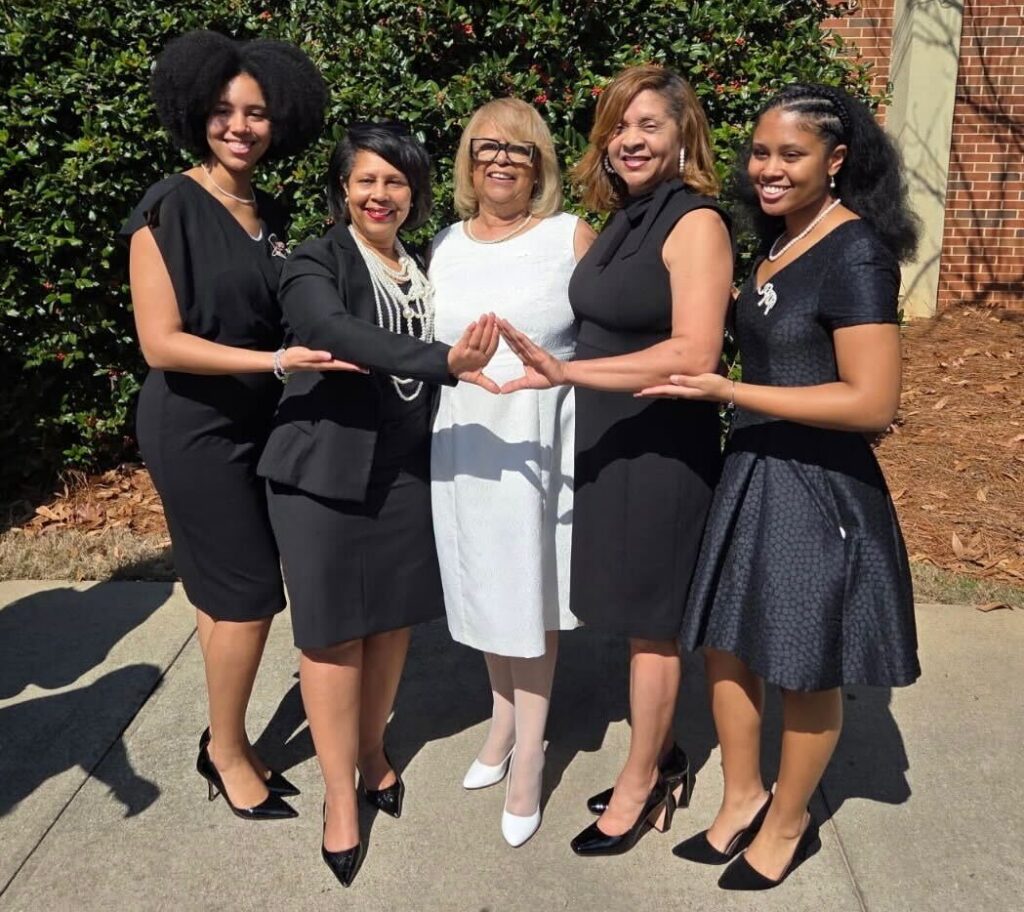 Five women stand outdoors in a row, four in black dresses and one in white, holding hands and smiling at the camera.