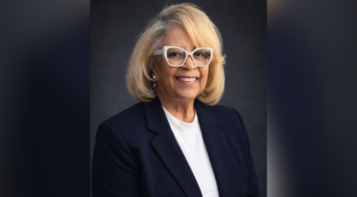 80-year-old works toward another milestone: A Mercer degree Smiling woman with blond hair, glasses, and black blazer poses against a dark background.