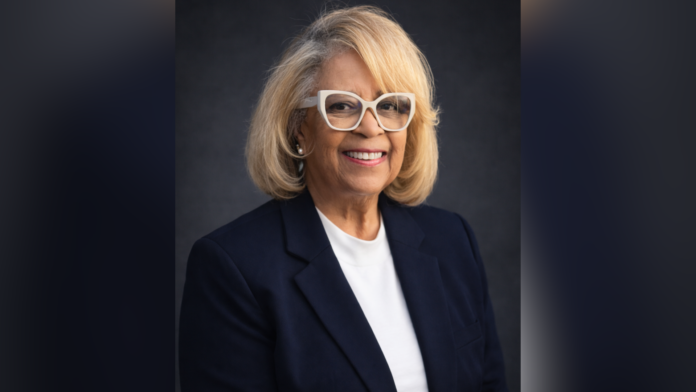 Smiling woman with blond hair, glasses, and black blazer poses against a dark background.