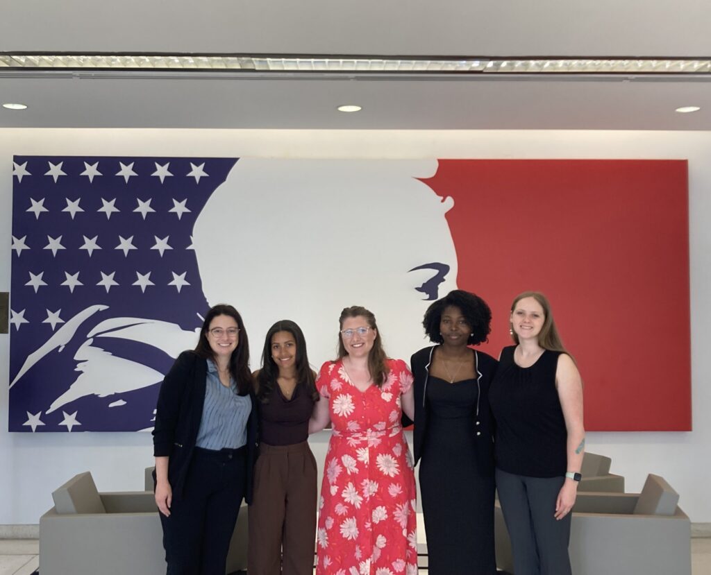 Five women standing together in front of a stylized artwork featuring the American flag and a white silhouette of a face.