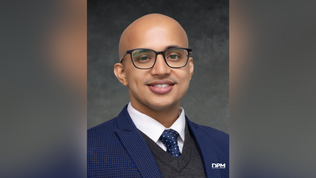 A man with glasses, a blue suit, and a polka dot tie smiles at the camera against a dark gray background.