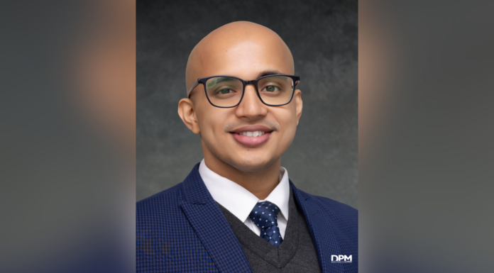 Dr.P.H. student selected for global health fellowship, public health think tank A man with glasses, a blue suit, and a polka dot tie smiles at the camera against a dark gray background.