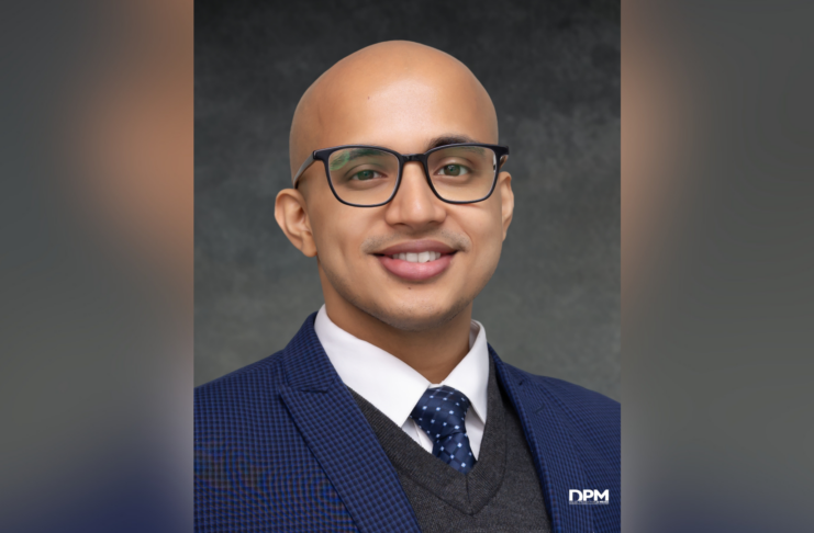 A man with glasses, a blue suit, and a polka dot tie smiles at the camera against a dark gray background.