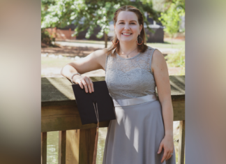 An ode to working adult students as they cross the finish line A woman in a gray dress stands outside on a wooden railing, holding a black folder and smiling at the camera.