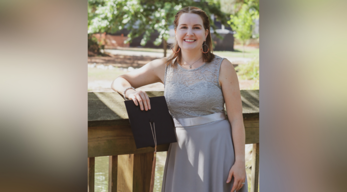 An ode to working adult students as they cross the finish line A woman in a gray dress stands outside on a wooden railing, holding a black folder and smiling at the camera.