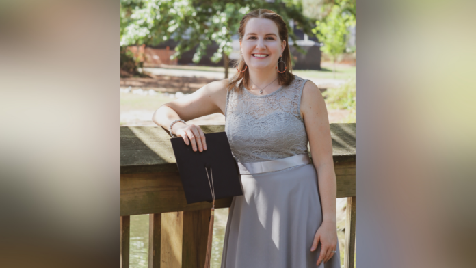A woman in a gray dress stands outside on a wooden railing, holding a black folder and smiling at the camera.