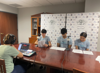Podcast explores Georgia’s role in Revolutionary War Three teenagers read scripts at a table while a person records audio using a laptop and mixer in a small studio.