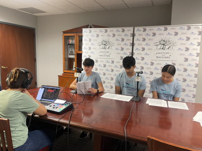 Three teenagers read scripts at a table while a person records audio using a laptop and mixer in a small studio.