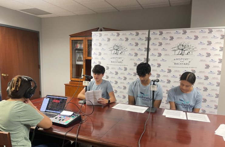 Three teenagers read scripts at a table while a person records audio using a laptop and mixer in a small studio.